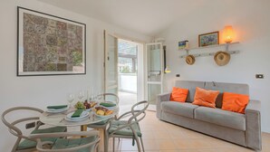 Un coin repas avec une table dressée pour deux personnes, un canapé gris agrémenté de coussins orange et une étagère murale où sont posés des chapeaux et une lampe.