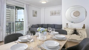 Un comedor con una mesa dispuesta para cuatro personas, un sofá gris, un mueble blanco con adornos y un amplio ventanal con vistas a edificios y palmeras.