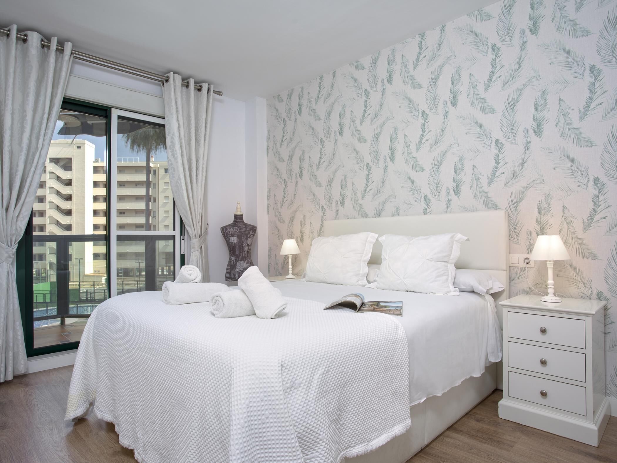 Ein Hotelzimmer mit einem großen Bett, weißer Bettwäsche, einem Nachttisch mit Lampe und einem Fenster mit Vorhängen.