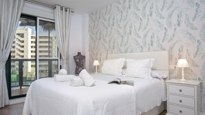 Una habitación de hotel con una cama grande, ropa de cama blanca, una mesita de noche con lámpara y una ventana con cortinas.