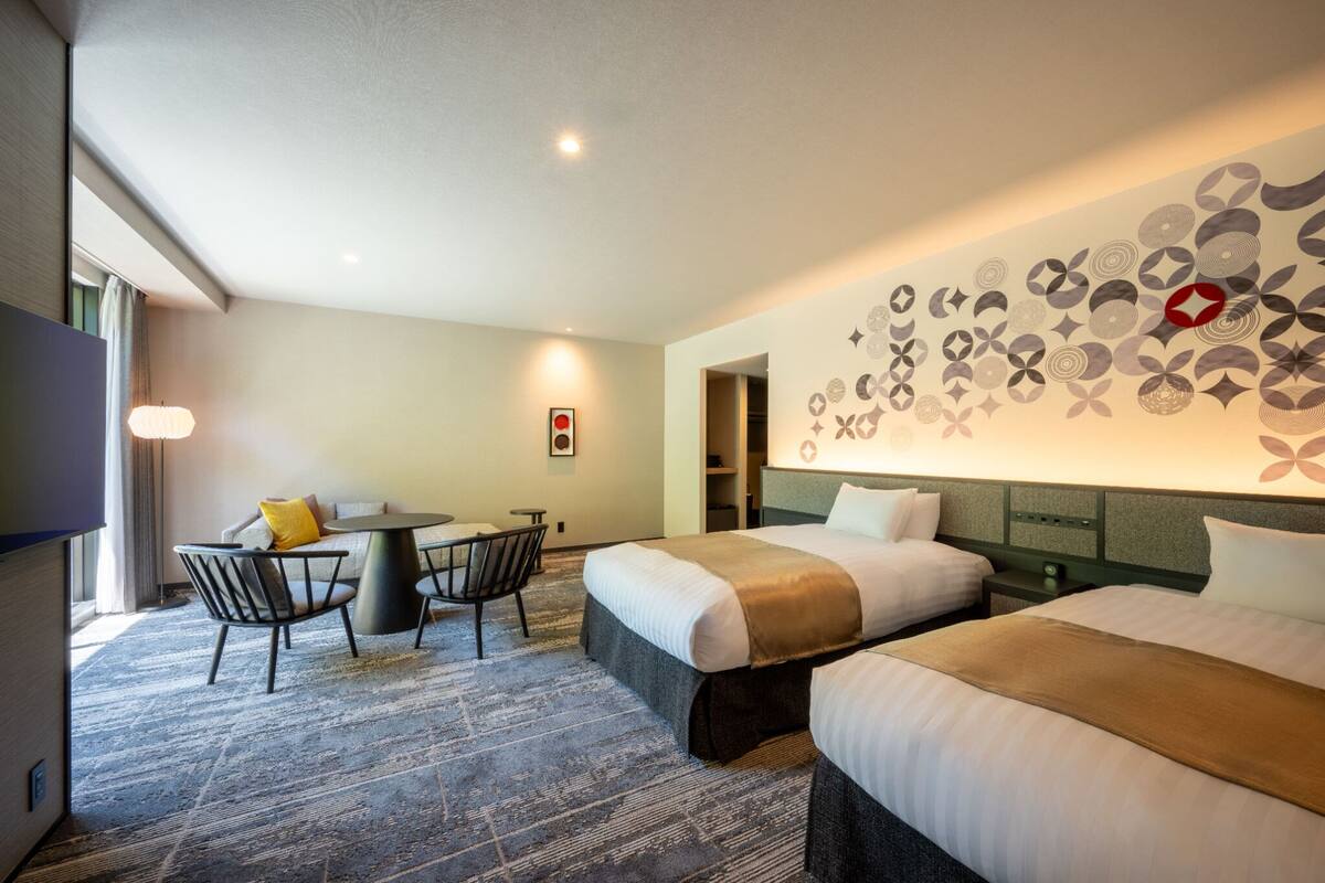 Quarto de hotel com duas camas, área de refeições e parede com padrão geométrico.