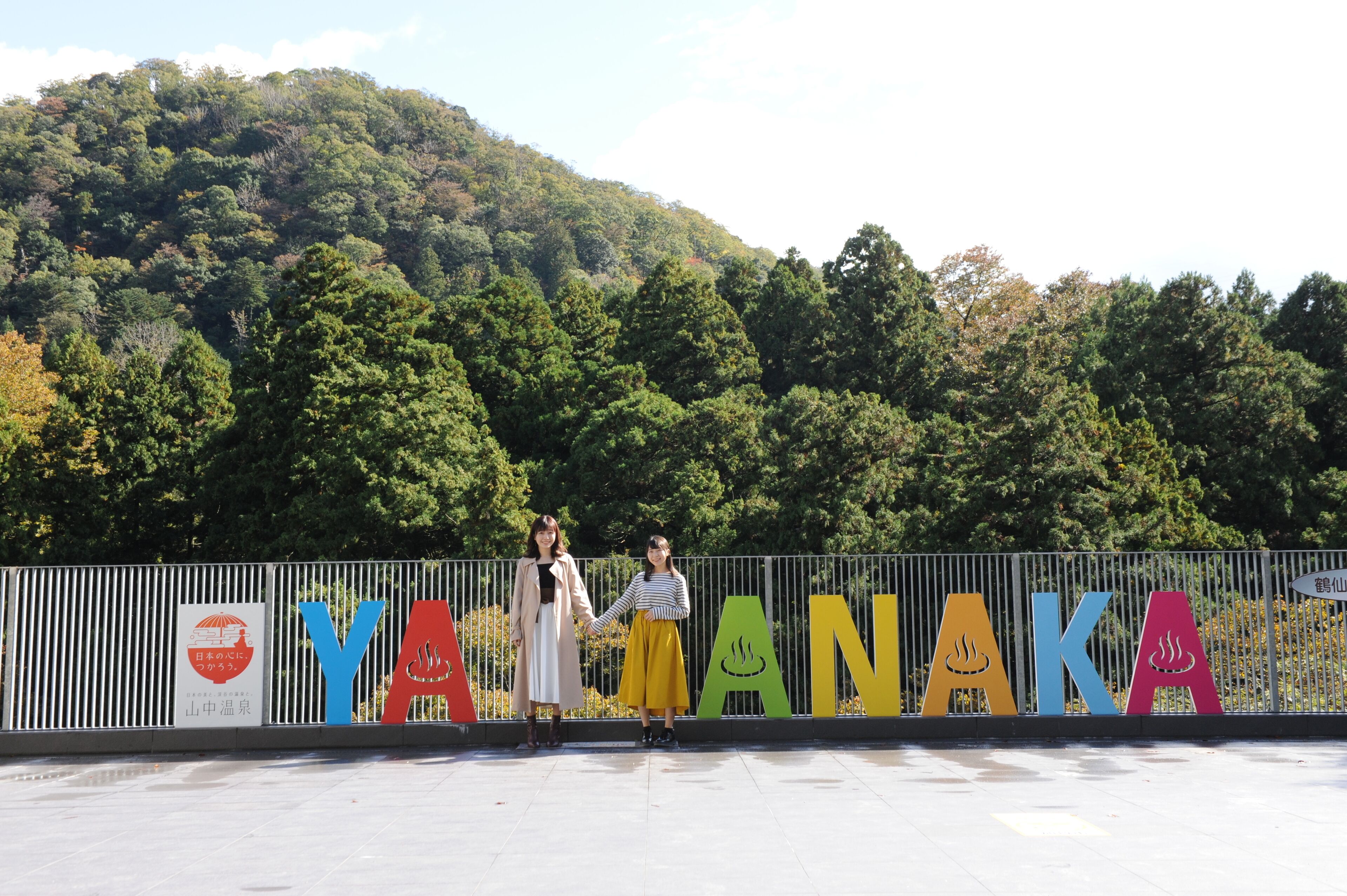 A sign with the word "YANAKA" in colorful letters, a railing, and a forested hill in the background.