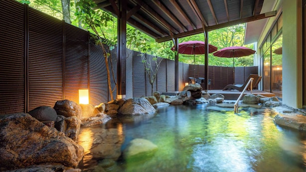 A serene indoor hot spring area with a wooden deck, large rocks, and a small waterfall. - Yamanaka Onsen KAJIKASO (Kaga)