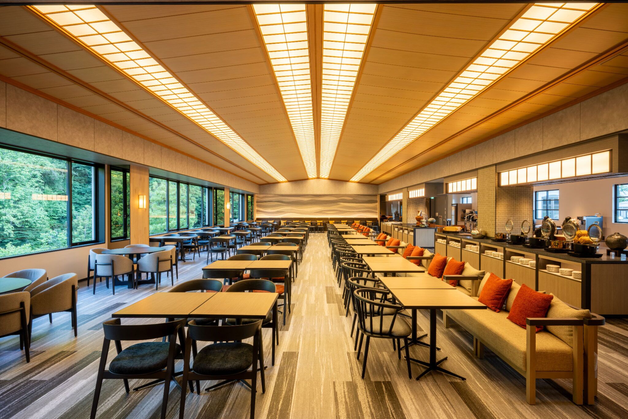 A spacious dining area with long tables and chairs, large windows offering views of greenery, and a modern ceiling design.