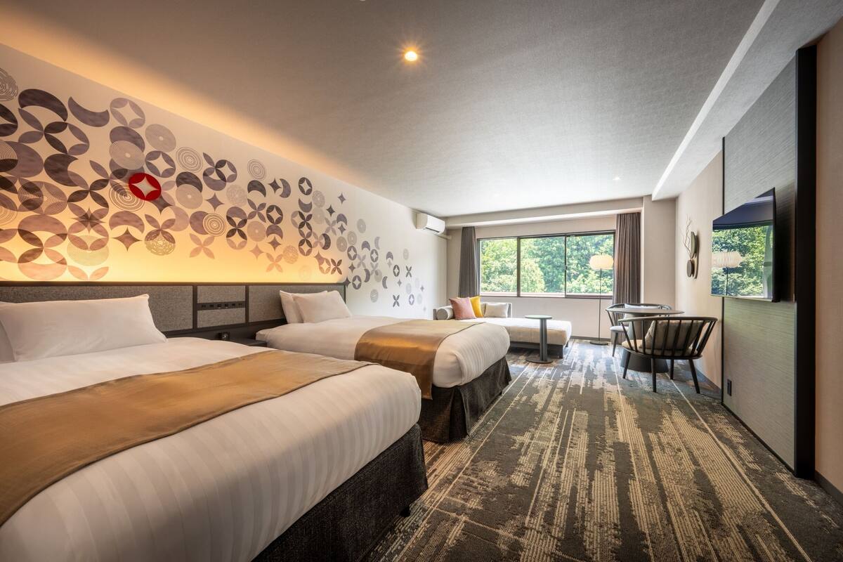 Quarto de hotel com duas camas, uma mesinha, uma cadeira, televisão e janela com vista para árvores.