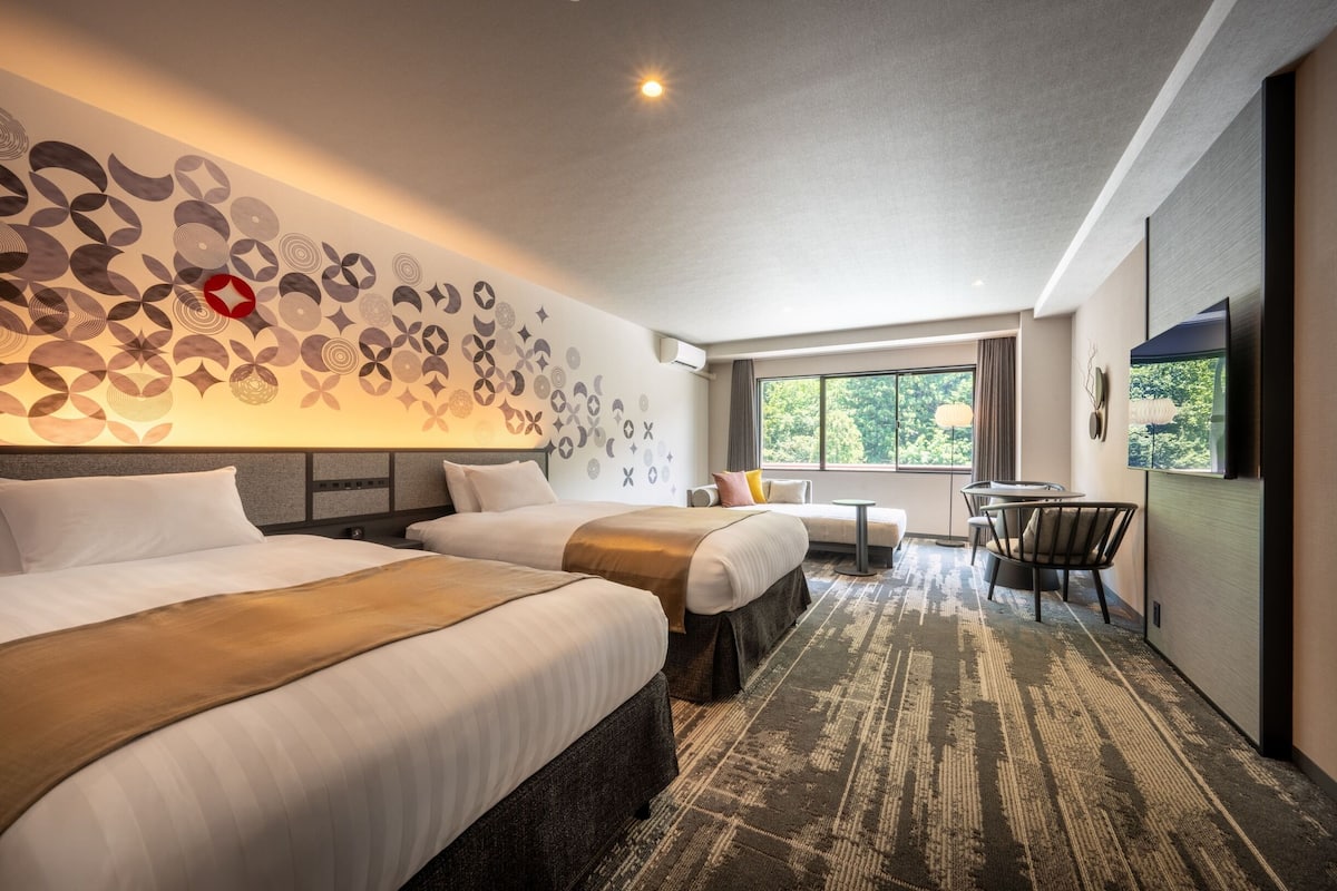 Habitación de hotel con dos camas, una mesita, una silla, televisión y ventana con vistas a árboles.