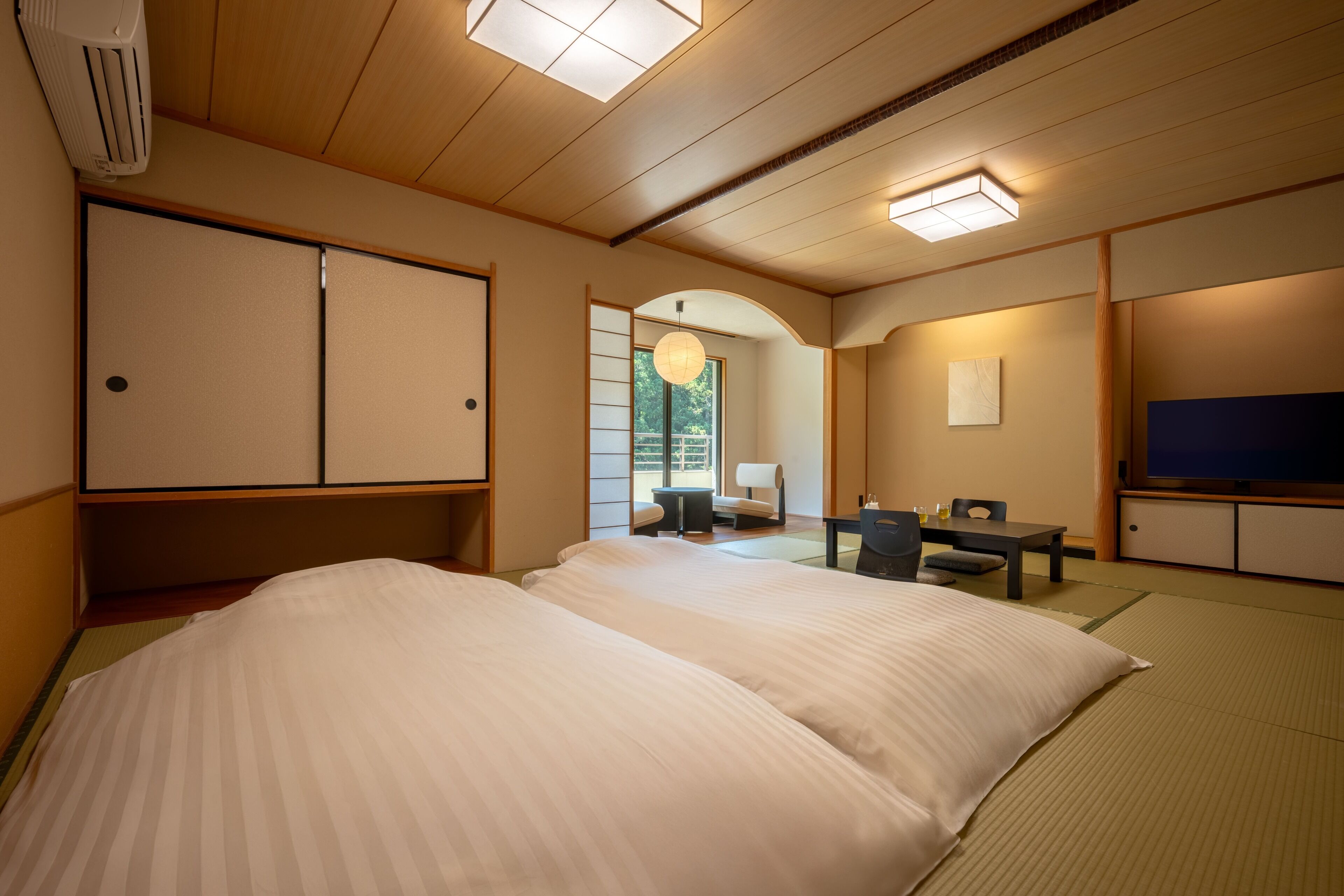 Una camera tradizionale in stile giapponese con pavimento in tatami, un tavolino con sedie, un letto con biancheria bianca, un televisore e una porta scorrevole.