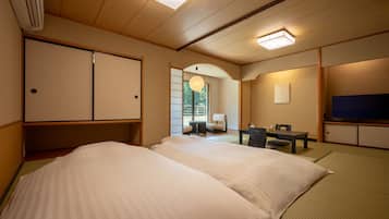 A traditional Japanese-style room with tatami flooring, a low table with chairs, a bed with white bedding, a television, and a sliding door.