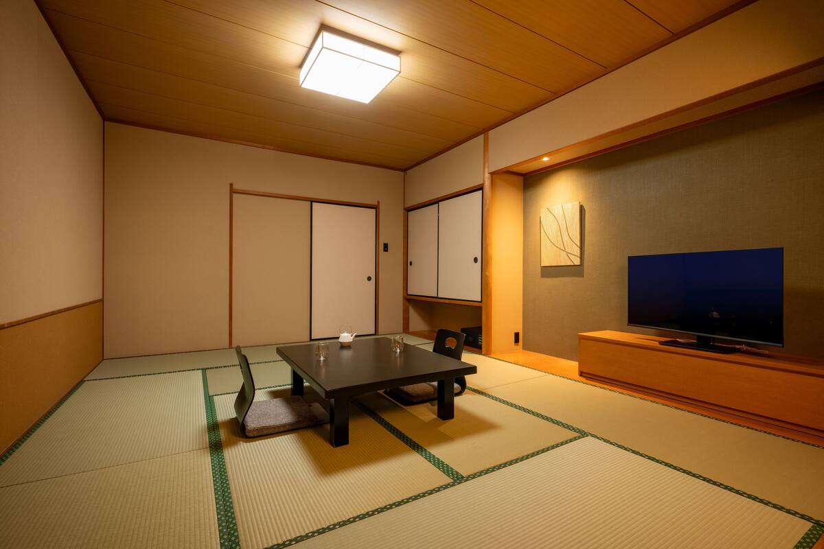 Um quarto japonês tradicional com tatames, uma mesa baixa, uma televisão e portas deslizantes.