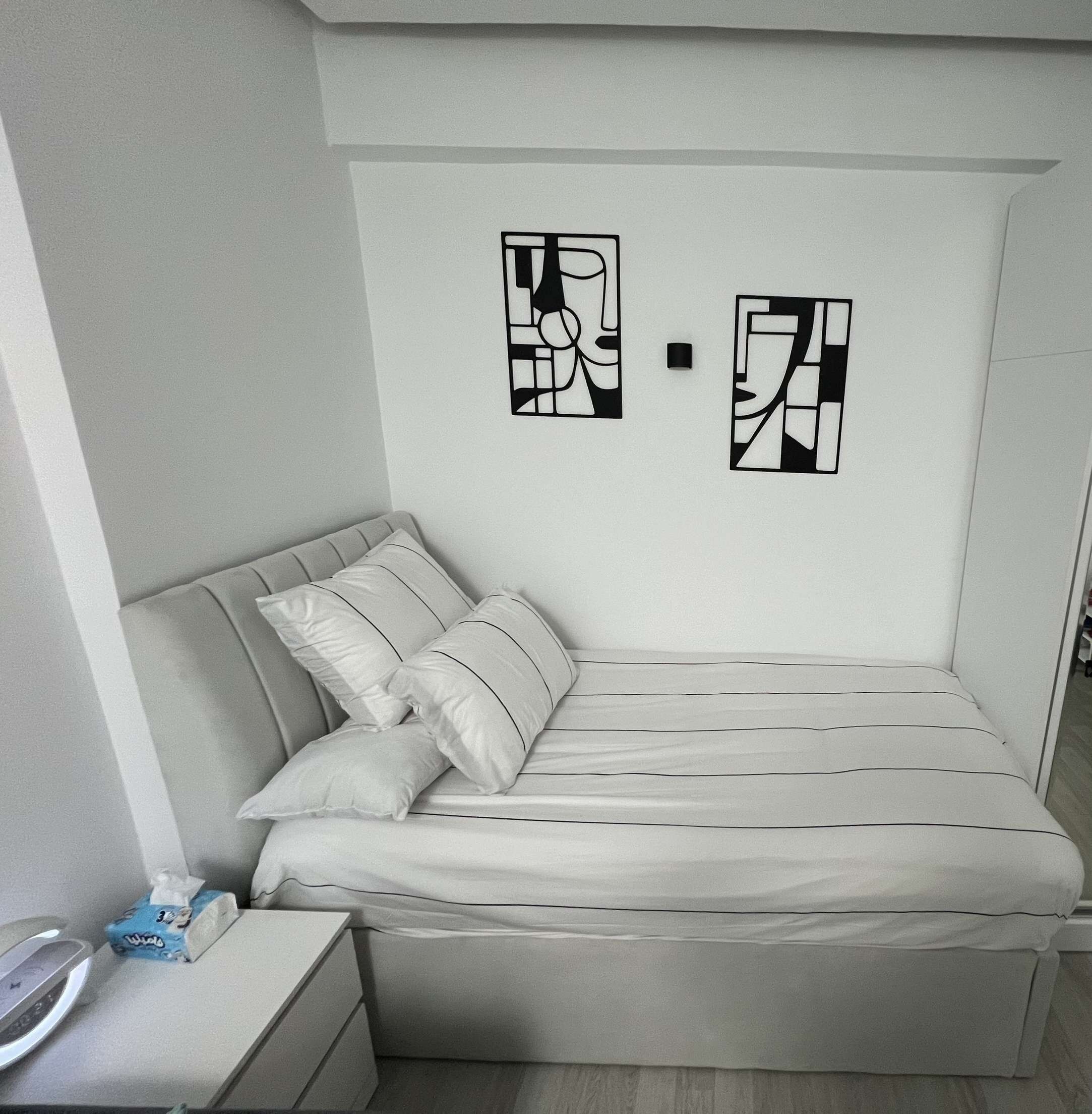Un dormitorio pequeño y ordenado con cama, almohadas, una mesita de noche y dos obras de arte en la pared.