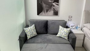 Une chambre moderne avec un canapé gris, deux coussins à motifs, une œuvre d’art représentant une sculpture classique accrochée au mur, et une table de chevet avec une lampe.