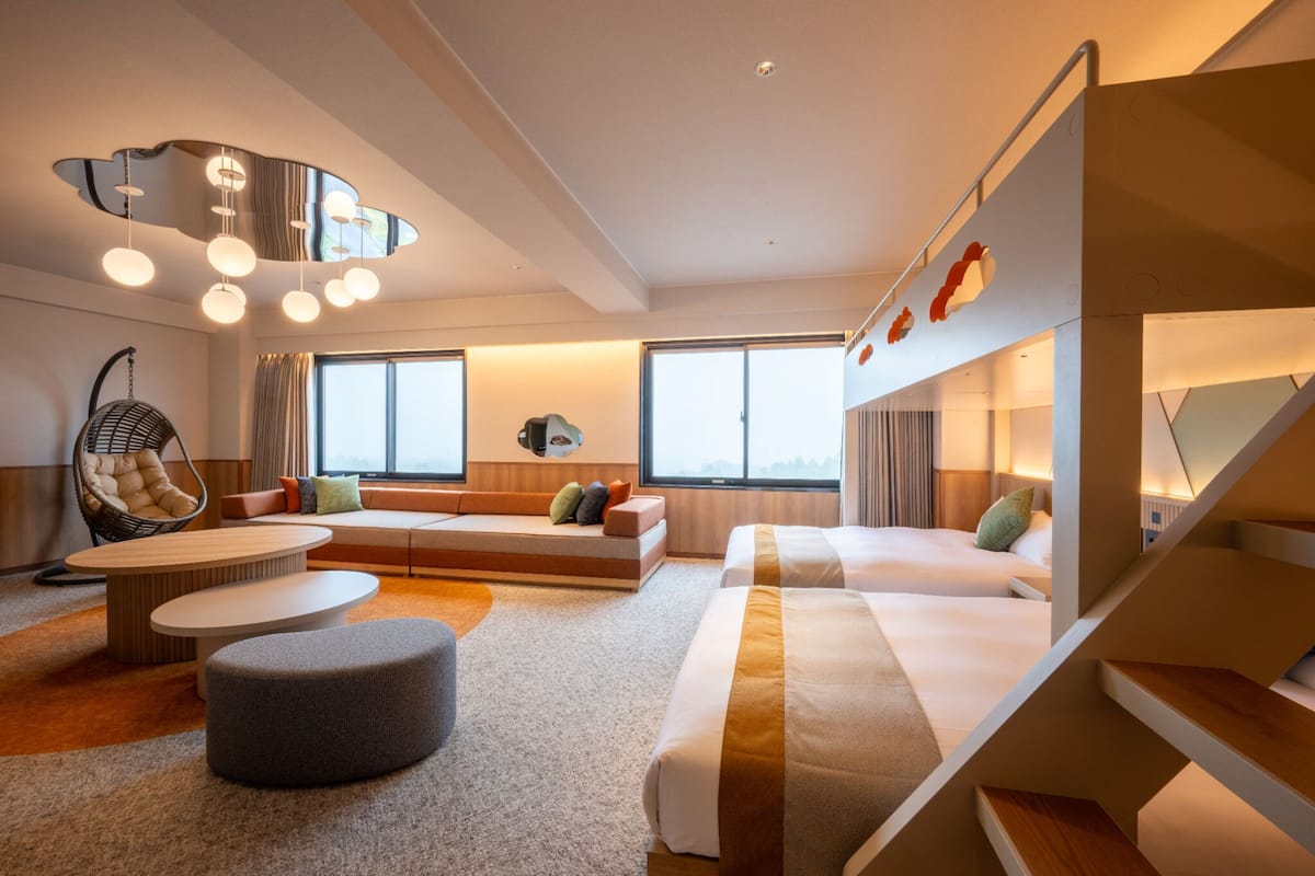 A modern hotel room with a sofa, ottoman, and a hanging chair. There is a staircase with a railing and a bed with pillows.