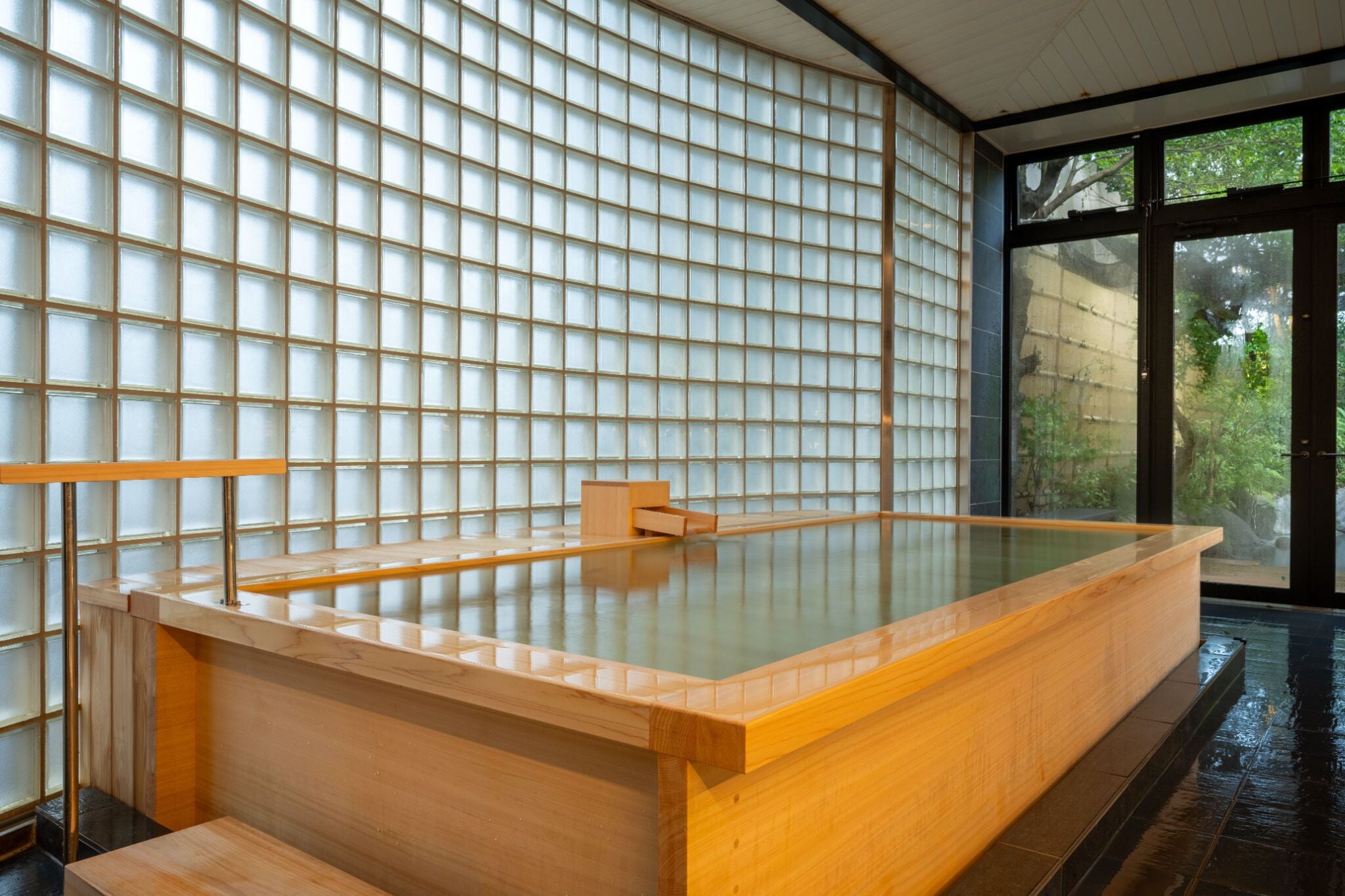 A traditional Japanese onsen with a wooden bath, tiled walls, and a view of greenery outside.