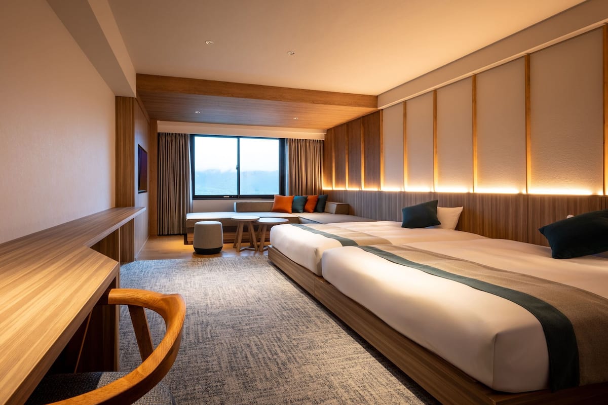 A hotel room with a large bed, a desk, a chair, a small table, and a view of the mountains through the window.