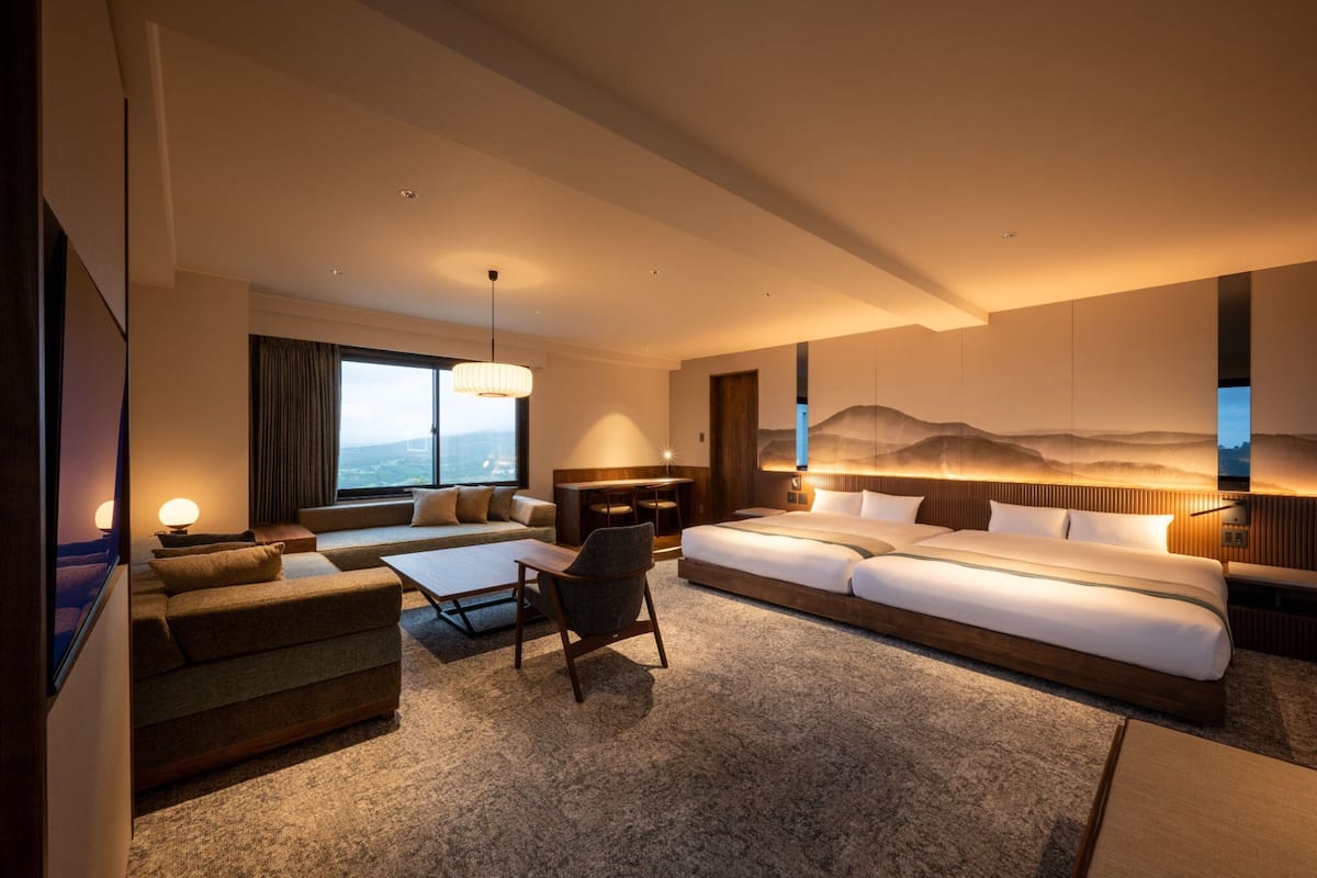 A modern hotel room with a large bed, a sofa, a desk, and a chair. There is a large window with curtains, a wall-mounted TV, and a painting of mountains.