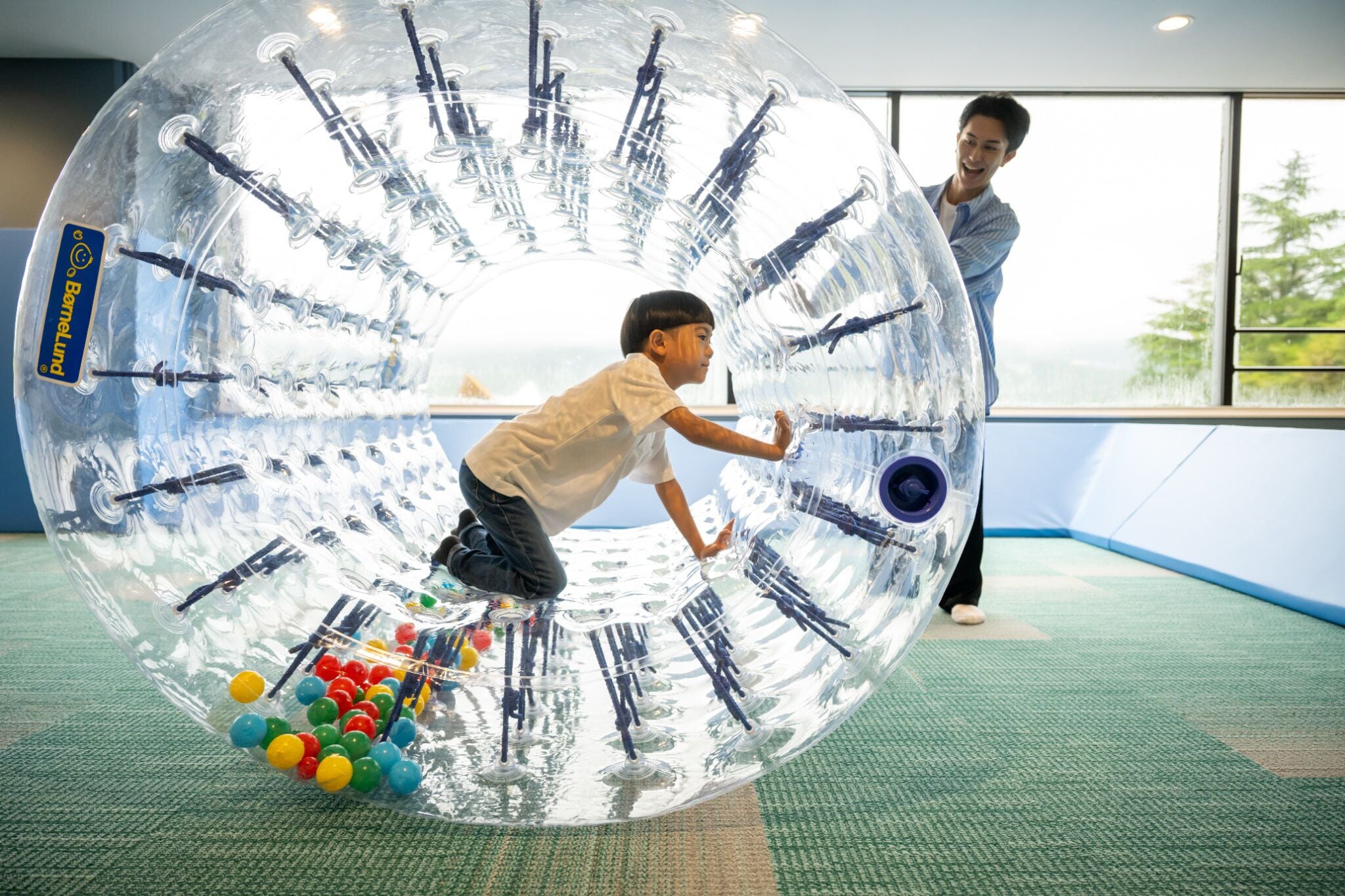 A transparent, spherical play structure with colorful balls inside, located in a room with large windows and a green carpet.