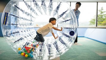 A transparent, spherical play structure with colorful balls inside, located in a room with large windows and a green carpet.