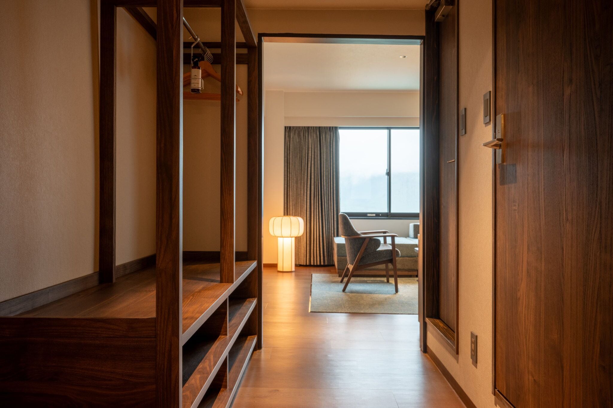 A hotel hallway with wooden flooring, a seating area with a lamp, and a view of a room with a large window.