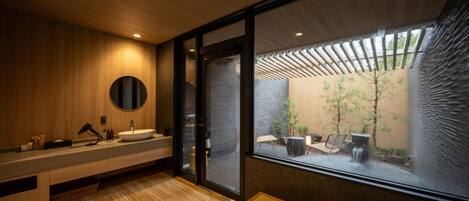 A modern bathroom with a wooden vanity, a round mirror, and a large window offering a view of a courtyard with plants and a seating area.