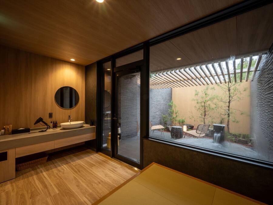 A modern bathroom with a wooden vanity, a round mirror, and a large window offering a view of a courtyard with plants and a seating area.