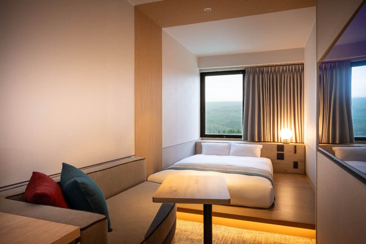 A hotel room with a bed, a small table, a sofa with cushions, a window with curtains, and a view of the landscape.