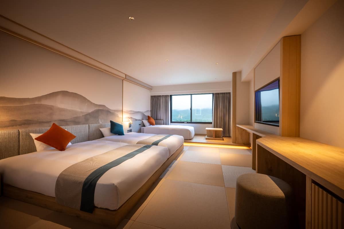 A hotel room with two beds, a TV, a desk, and a large window.