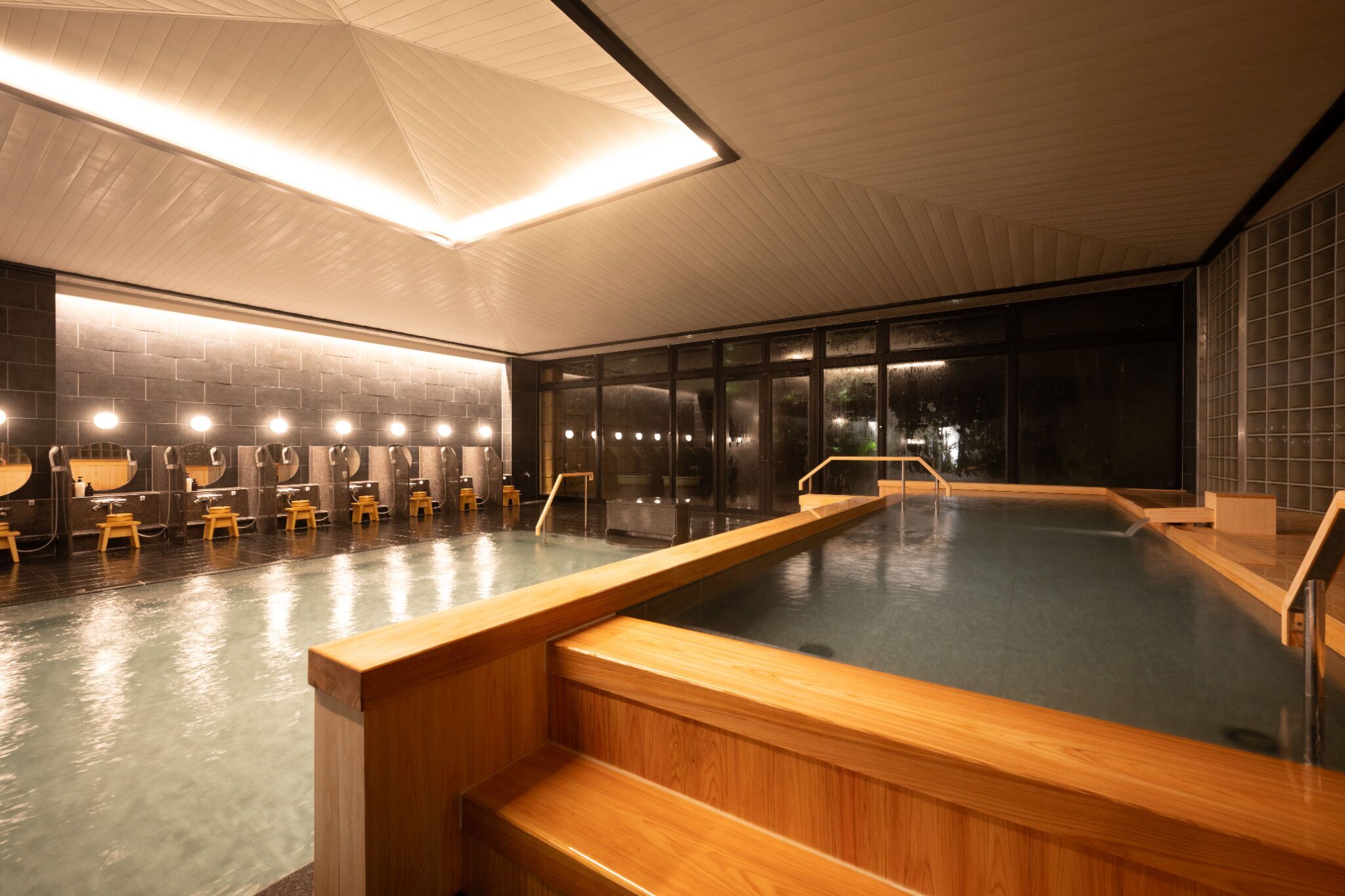 A modern indoor swimming pool with wooden steps leading into the water, a brick wall with built-in benches, and a large skylight above.