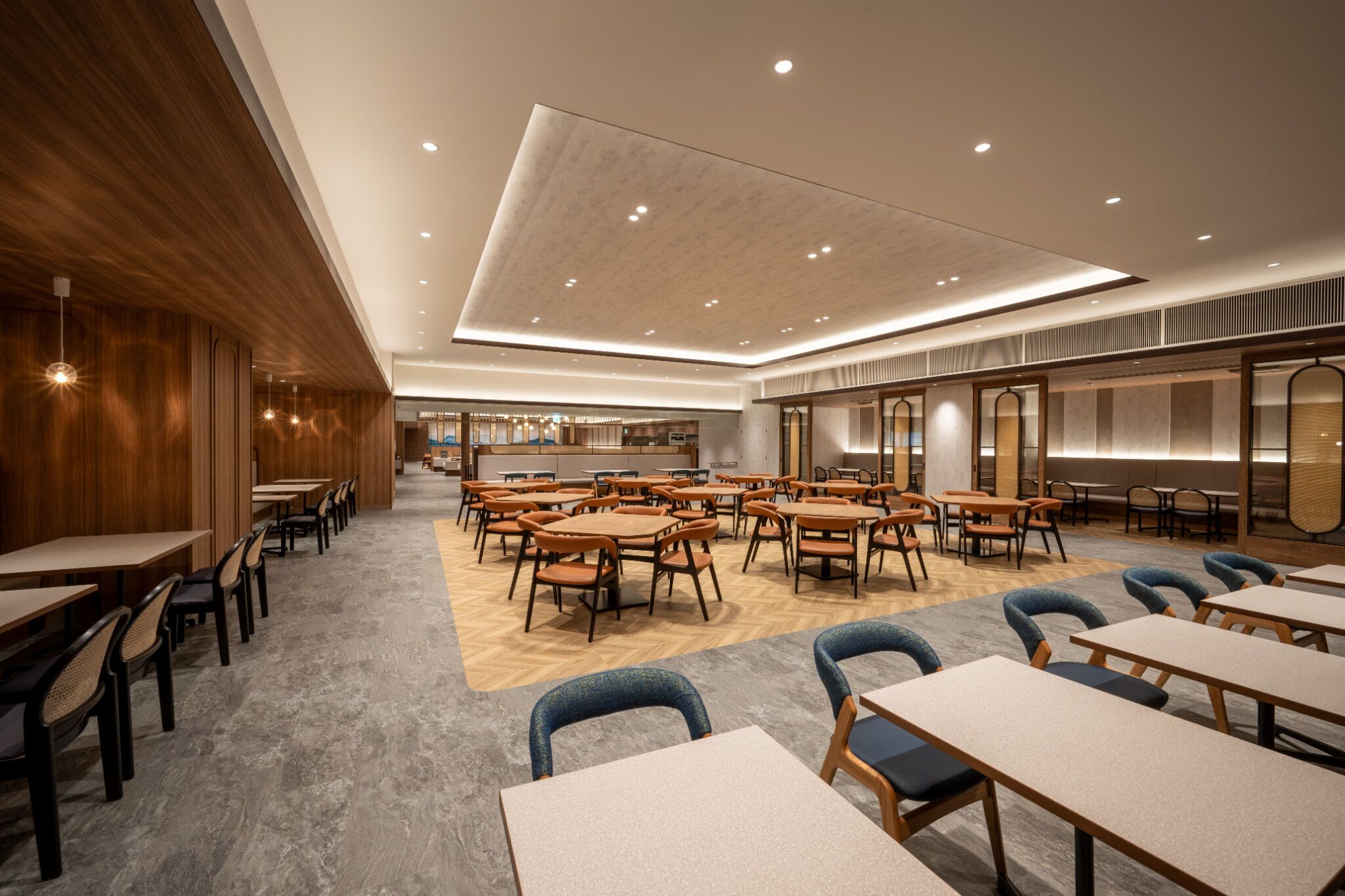 A spacious hotel dining area with multiple tables and chairs, wooden paneling on one side, and a modern ceiling design.