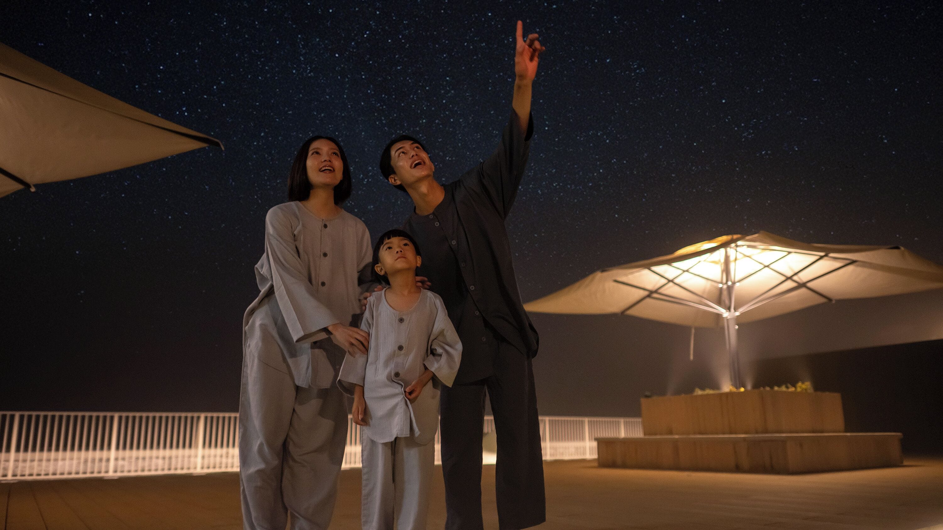 Una familia de tres bajo un cielo estrellado, con una gran estructura tipo sombrilla al fondo.