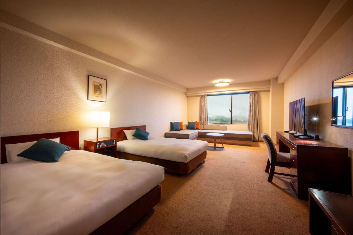 A hotel room with two beds, a desk, a chair, a lamp, and a window with curtains.