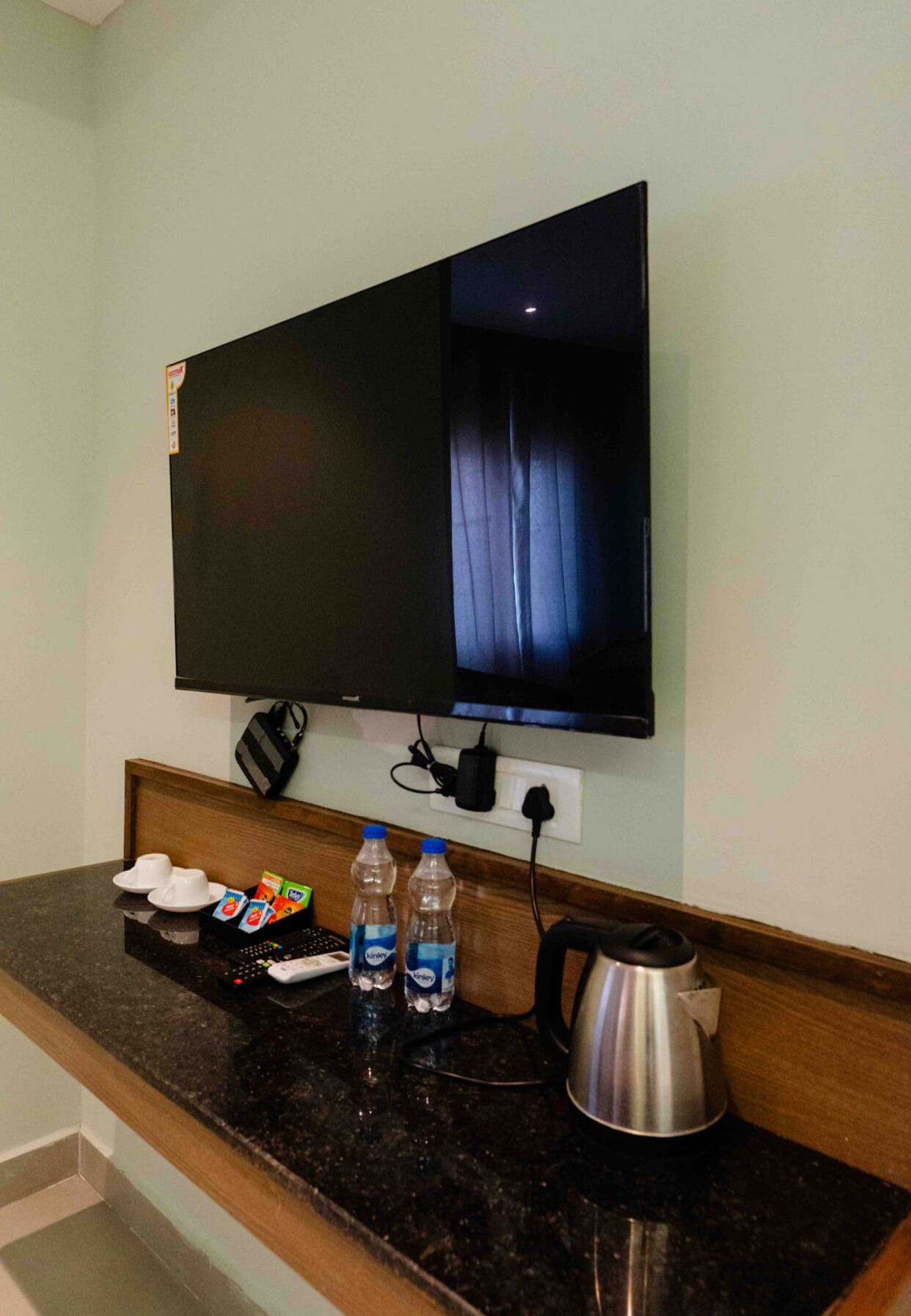 Habitación de hotel con televisor de pantalla plana montado en la pared, una encimera de madera con una tetera, dos botellas de agua, un control remoto y una bandeja con tazas y un paquete de aperitivos.