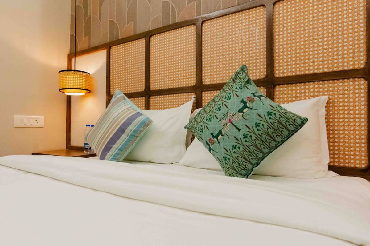 Un lit bien fait, avec du linge de lit blanc et des coussins décoratifs, une tête de lit en osier et une lampe fixée au mur.