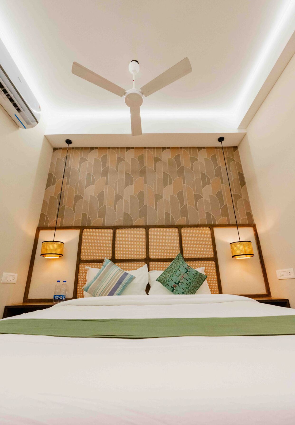 Une chambre d’hôtel avec un lit, un ventilateur de plafond, deux lampes fixées au mur et une tête de lit à motifs.
