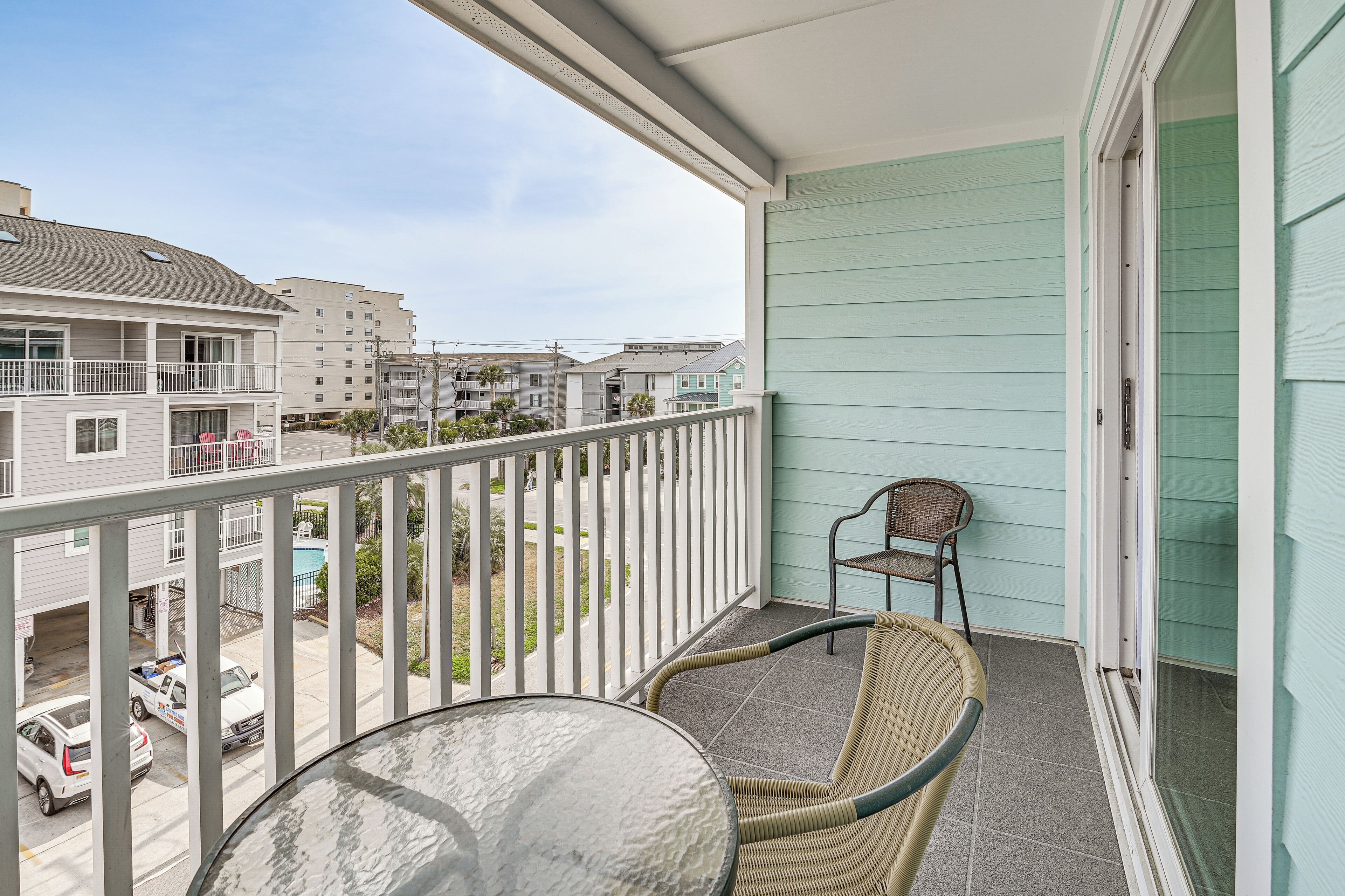 A balcony with a wicker chair and a table, overlooking a residential area with houses and a parking lot.