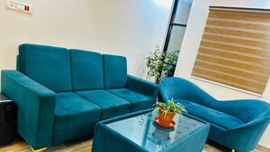 Eine Sitzgruppe mit einem türkisfarbenen Sofa und Sesseln, einem Glastisch, einer Topfpflanze und einer Steckdose an der Wand.