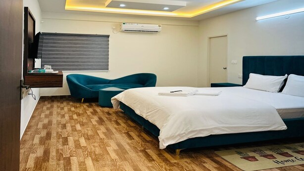 Ein modernes Hotelzimmer mit Holzboden, einem Bett mit weißer Bettwäsche, einem saphirfarbenen Sessel, einem kleinen runden Tisch und einem Fernseher auf einem Holzbodenregal.