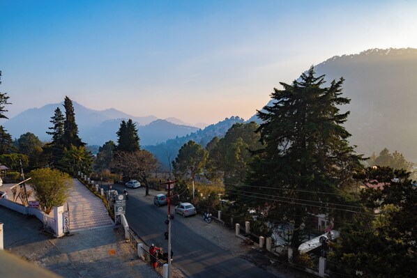 A road leading to a residential area with houses and trees, surrounded by mountains. - Dew Drops Homestay (Nainital)