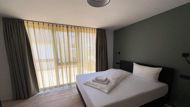 A hotel room with a large bed, wooden flooring, a window with curtains, and a ceiling light. - New Age Boardinghaus Bad Rappenau (Bad Rappenau)