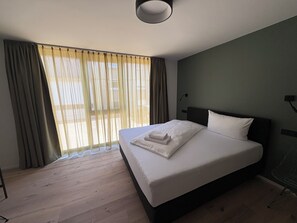 A hotel room with a large bed, wooden flooring, a window with curtains, and a ceiling light. - New Age Boardinghaus Bad Rappenau (Bad Rappenau)