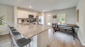 Una cocina moderna con encimera de granito, armarios blancos y horno empotrado. Cuenta con una zona de comedor con banco y mesa.