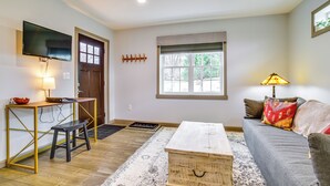 Un salon comprenant un canapé, une table basse, un téléviseur, un banc en bois et une fenêtre avec des stores.