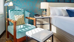 A hotel room with a bed, a wooden chair with a patterned cushion, a small table, and a nightstand with a lamp.