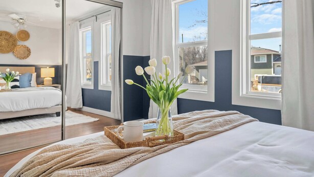 A bedroom with a bed, a bedside table with a lamp, a window with curtains, and a mirror reflecting the room. - Bluebird | Charming 3-Bedroom Home in Drumheller! (Drumheller)