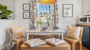 A dining area with a white table, two wooden chairs, a woven pendant light, and a window with floral curtains. - Bluebird | Charming 3-Bedroom Home in Drumheller! (Drumheller)