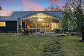 Una casa moderna con un amplio ventanal de vidrio, un comedor con sillas y un sendero de piedra que lleva a la entrada.