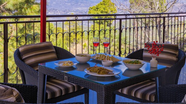 A dining table with plates of food and glasses of red wine, surrounded by wicker chairs with striped cushions, set on a balcony with a view of mountains and trees. - Alaya Stays Captains Nest 3 BHK Retreat With Garden & Scenic Views (Kasauli)