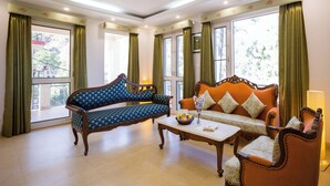 A spacious living room with a blue patterned sofa, an orange sofa with floral cushions, a wooden coffee table, and large windows with green curtains. - Alaya Stays Captains Nest 3 BHK Retreat With Garden & Scenic Views (Kasauli)