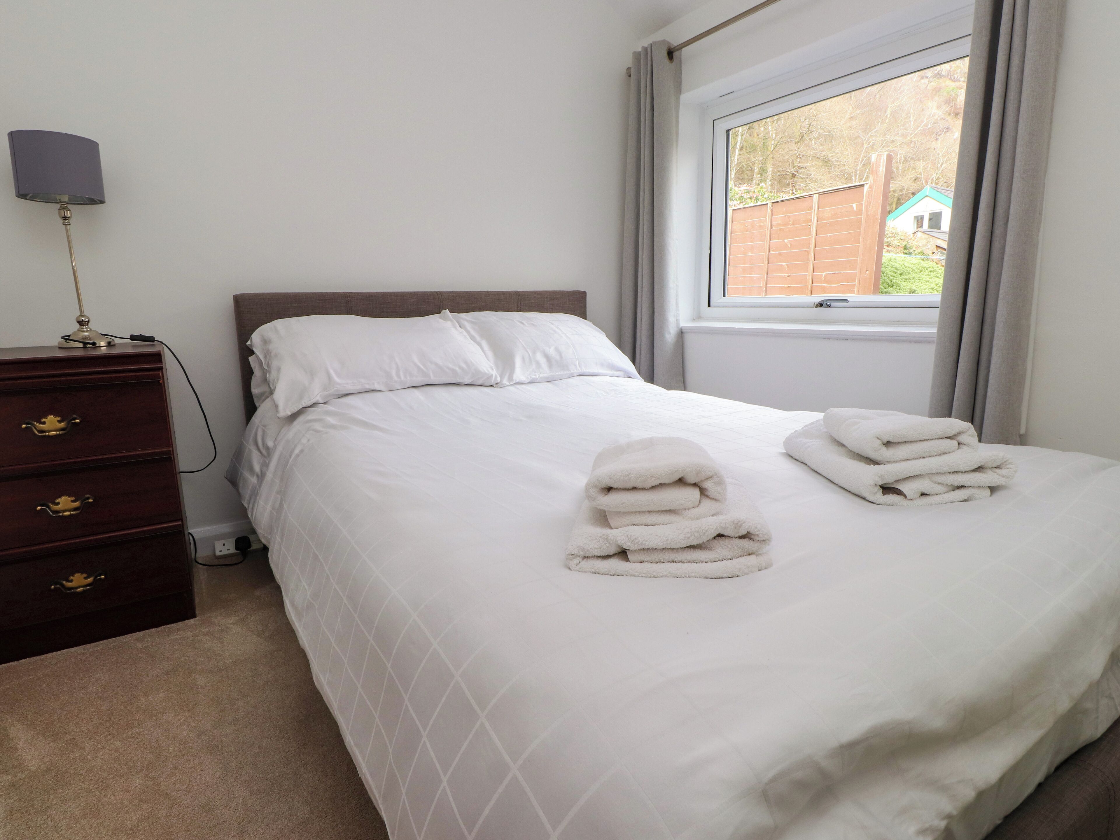 A neatly made bed with white linens and towels, a wooden dresser with drawers, a lamp, and a window with curtains.