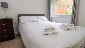 A neatly made bed with white linens and towels, a wooden dresser with drawers, a lamp, and a window with curtains.