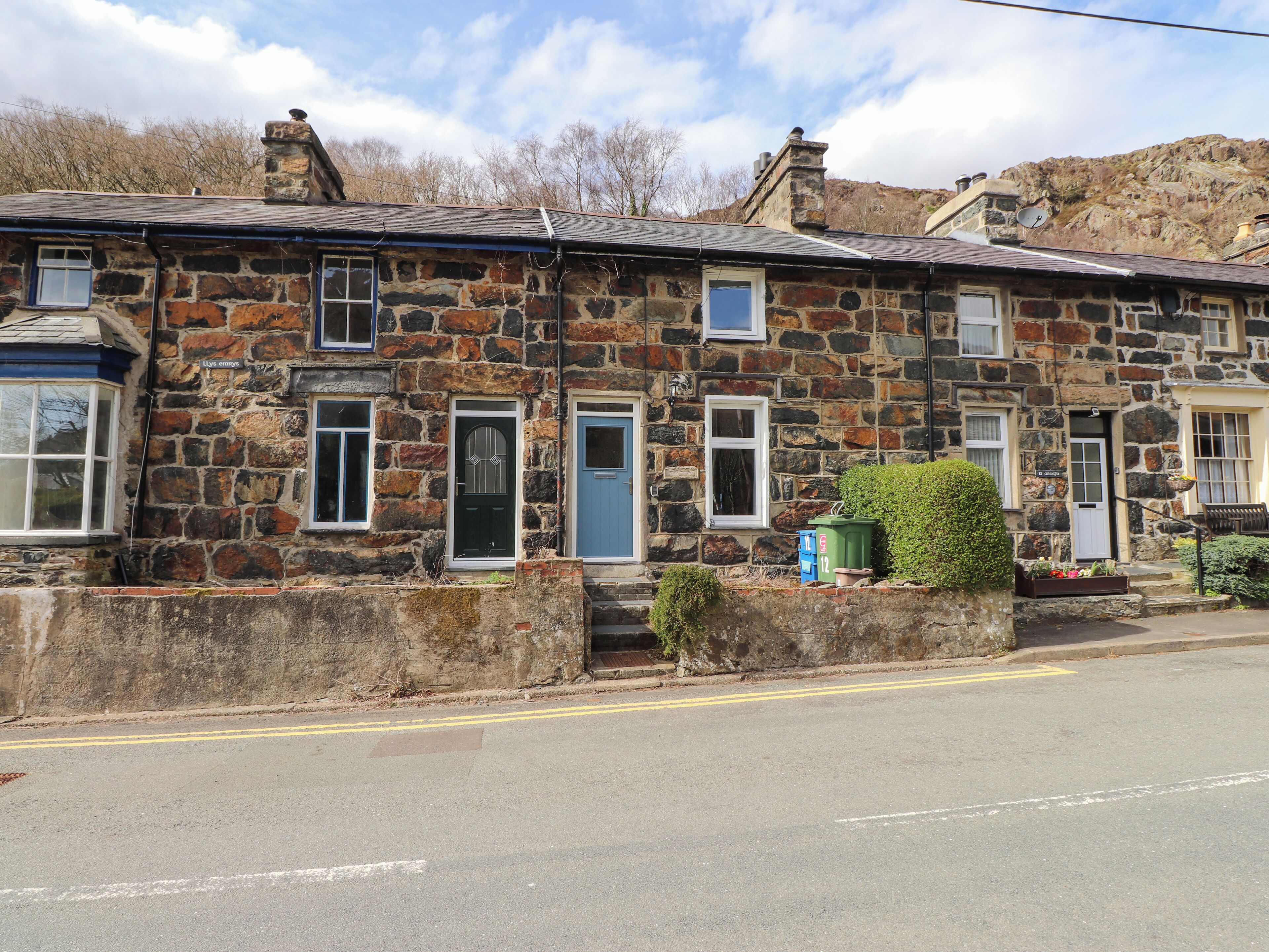 A stone building with multiple doors and windows, a small garden area with a bench, and a clear road in front.